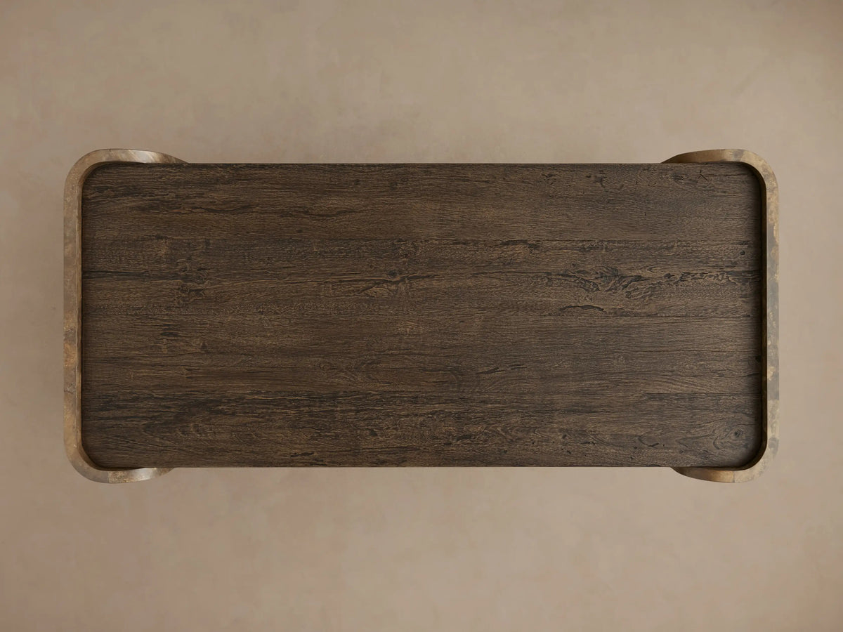Close-up of Alvaro Coffee Table. Dark brown spalted primavera wood top & shelf with grain, connected by curved weathered aluminum panel with mottled bronze/gold patina.