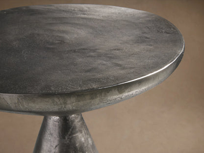 Amina End Table in splash nickel finish, showing a sleek, reflective surface with a modern, hourglass shape on a wooden floor.