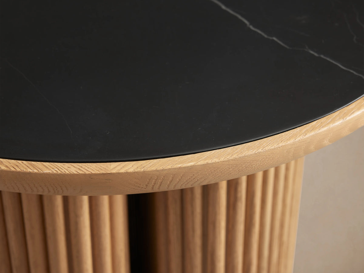 Astor Round End Table with a black marble top and cylindrical oak reed base on a wooden floor, showcasing artisan craftsmanship.