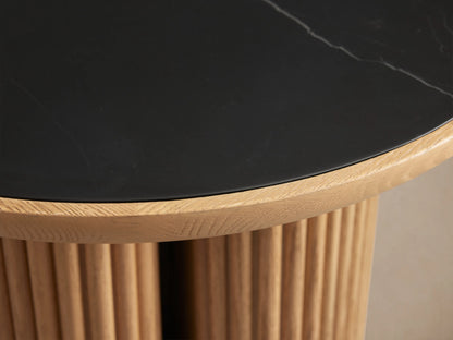Astor Round End Table with a black marble top and cylindrical oak reed base on a wooden floor, showcasing artisan craftsmanship.