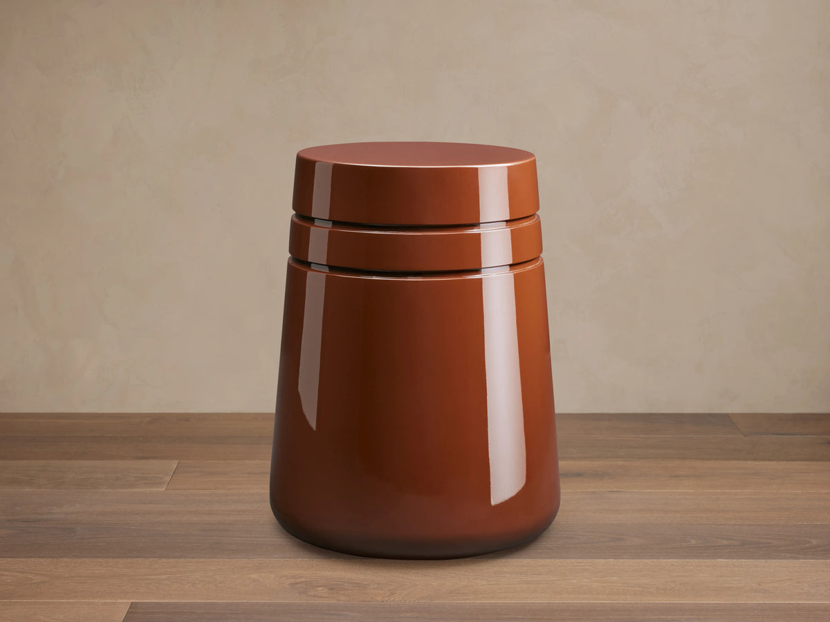 August Lacquer Stool with a high-gloss finish in a warm brown color, featuring a smooth, tapered cylindrical shape on a wooden floor.