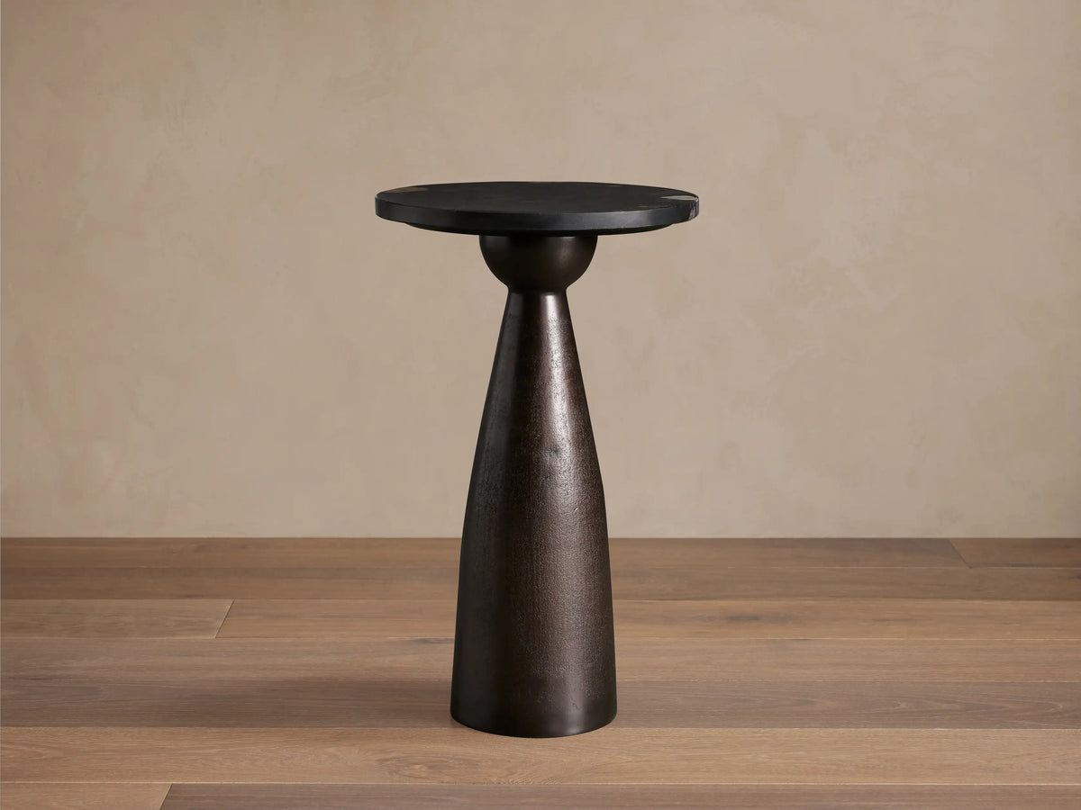 Beaufort Outdoor Martini Table with a dark mottled bronze conical base and a black round top, set on a textured stone floor.