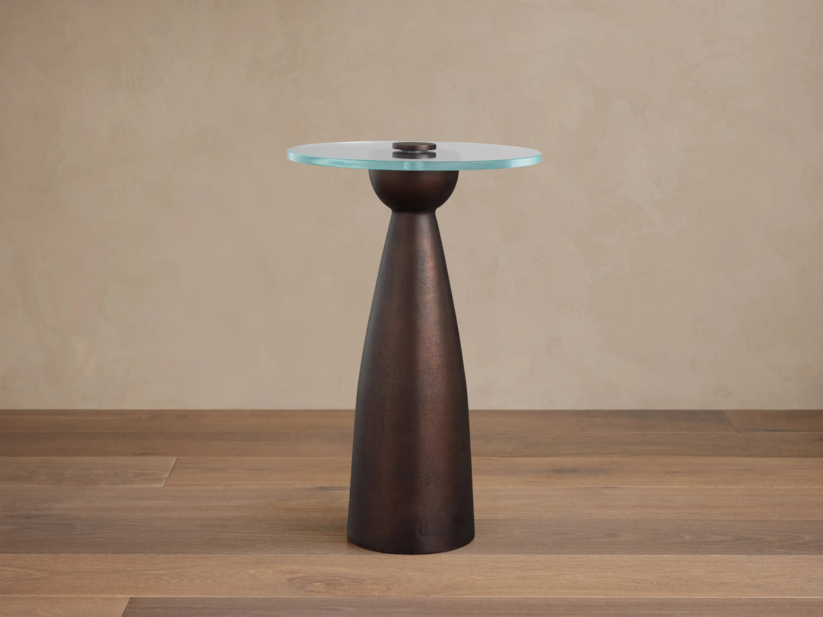 Beaufort Glass Top Martini Table with a round clear glass top and conical dark metallic bronze base on a textured stone floor.