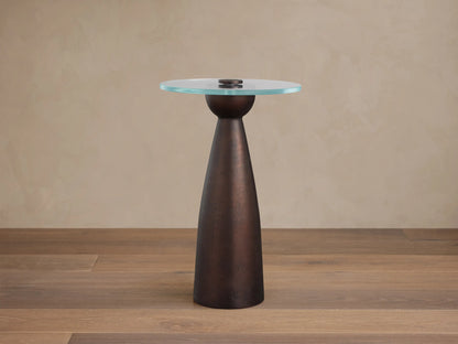 Beaufort Glass Top Martini Table with a round clear glass top and conical dark metallic bronze base on a textured stone floor.