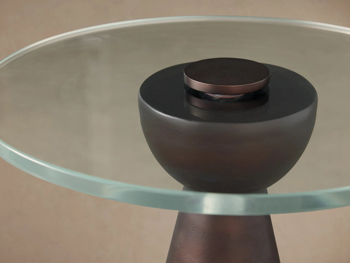 Beaufort Glass Top Martini Table with a dark, tapered bronze-finished base and a clear, round glass top, on a wooden floor.