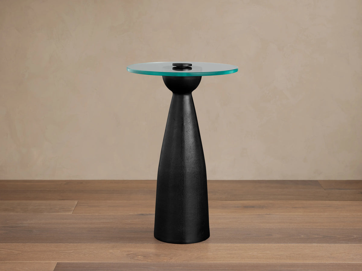 Beaufort Glass Top Martini Table with a round, clear glass tabletop and a conical black cast aluminum base on a wooden floor.