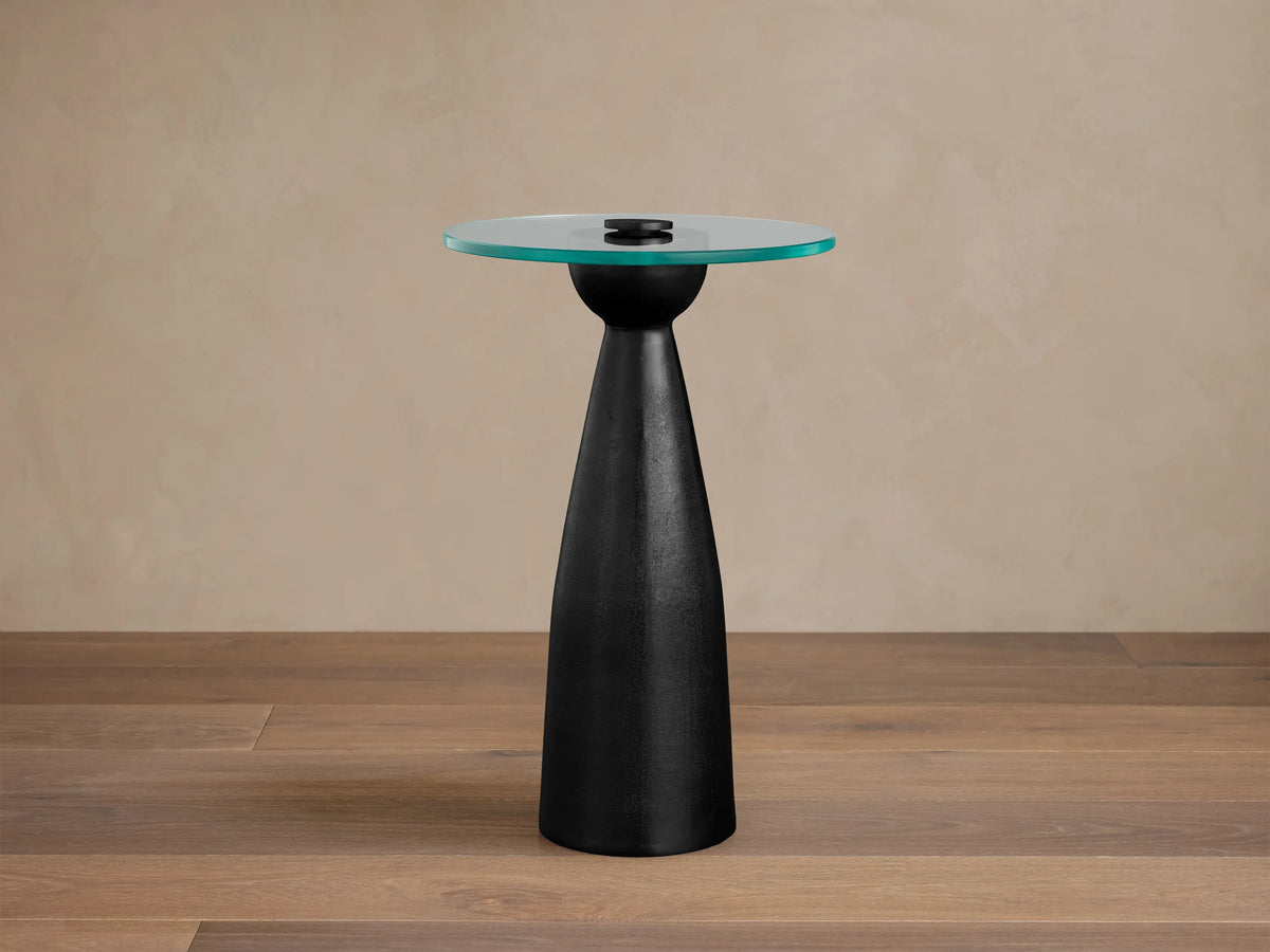 Beaufort Glass Top Martini Table, round glass top with faint green tint, on a dark, tapered pedestal base. Set on light stone floor.