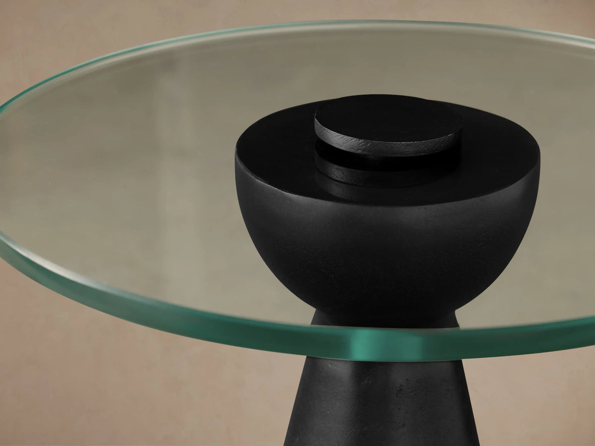 Beaufort Glass Top Martini Table: round clear glass top on a textured black tapered base, against a beige wall & wood floor.
