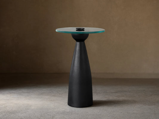 Beaufort Glass Top Martini Table, round glass top with faint green tint, on a dark, tapered pedestal base. Set on light stone floor.