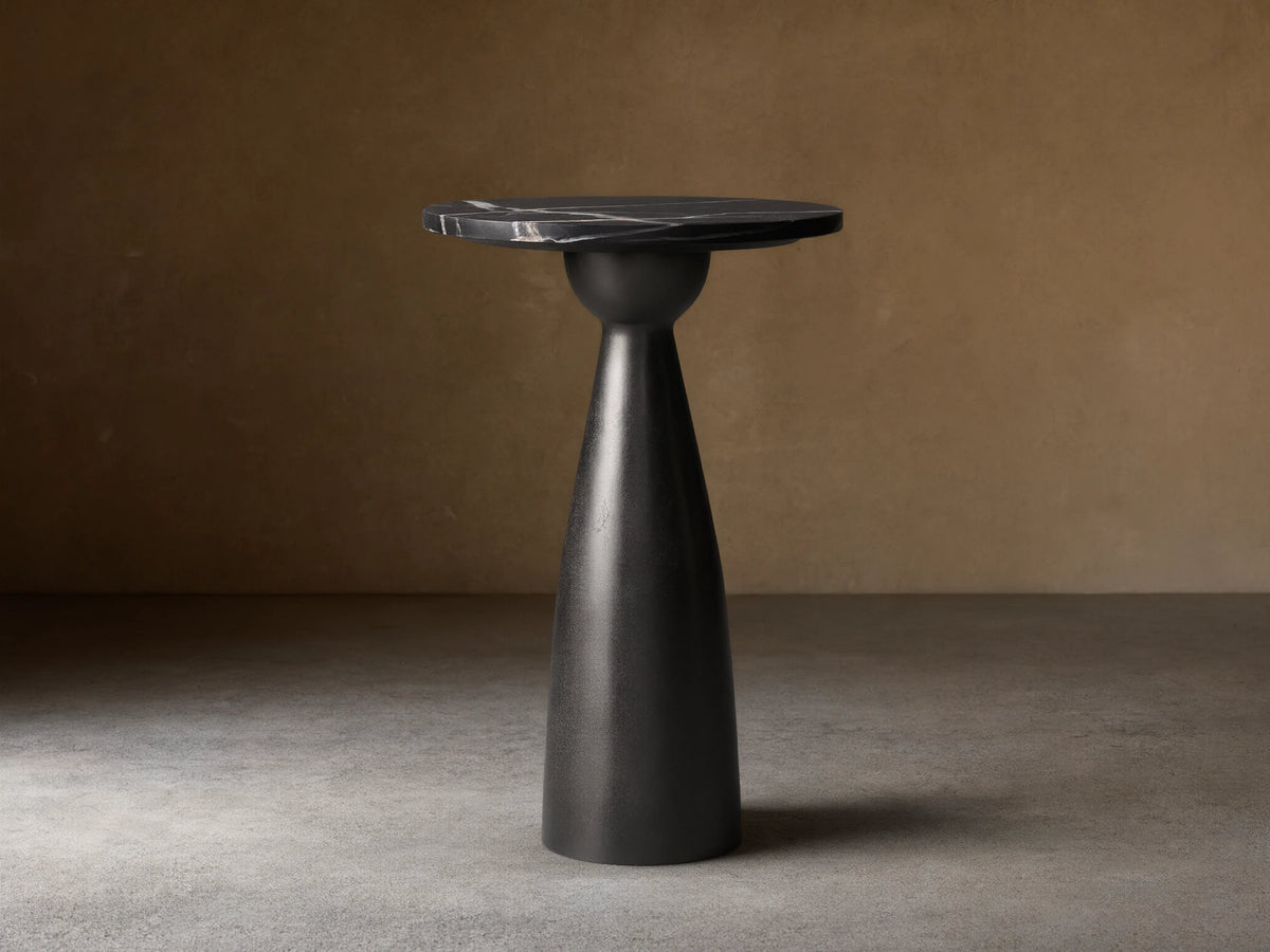 Beaufort Martini Table: round black marble top with white veining on a dark, textured conical base, sitting on stone tiles.
