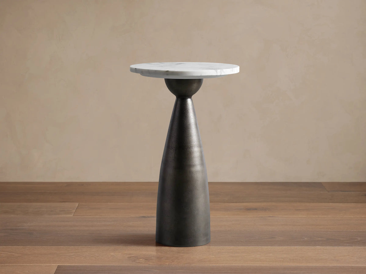 Beaufort Martini Table: round white marble top, dark textured conical base, on light stone floor against a beige wall.