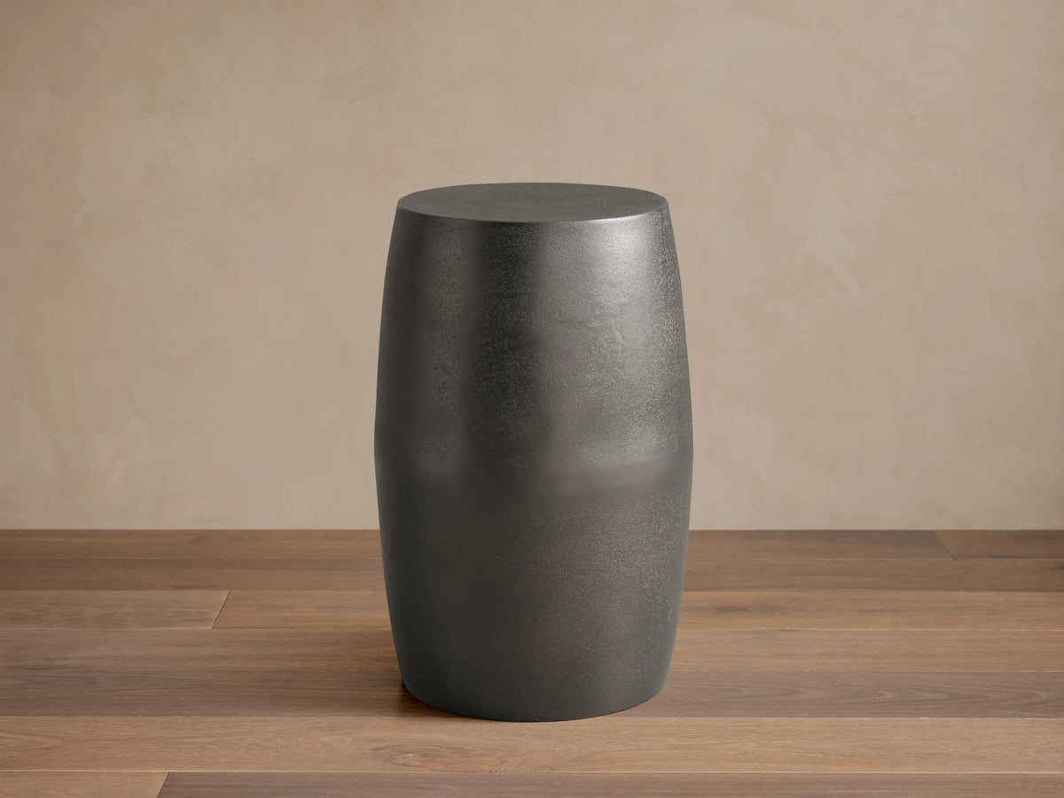 Antiqued dark gray Hammered Drum End Table with a textured finish, standing on a tiled floor against a light beige wall.