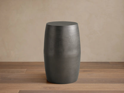 Antiqued dark gray Hammered Drum End Table with a textured finish, standing on a tiled floor against a light beige wall.