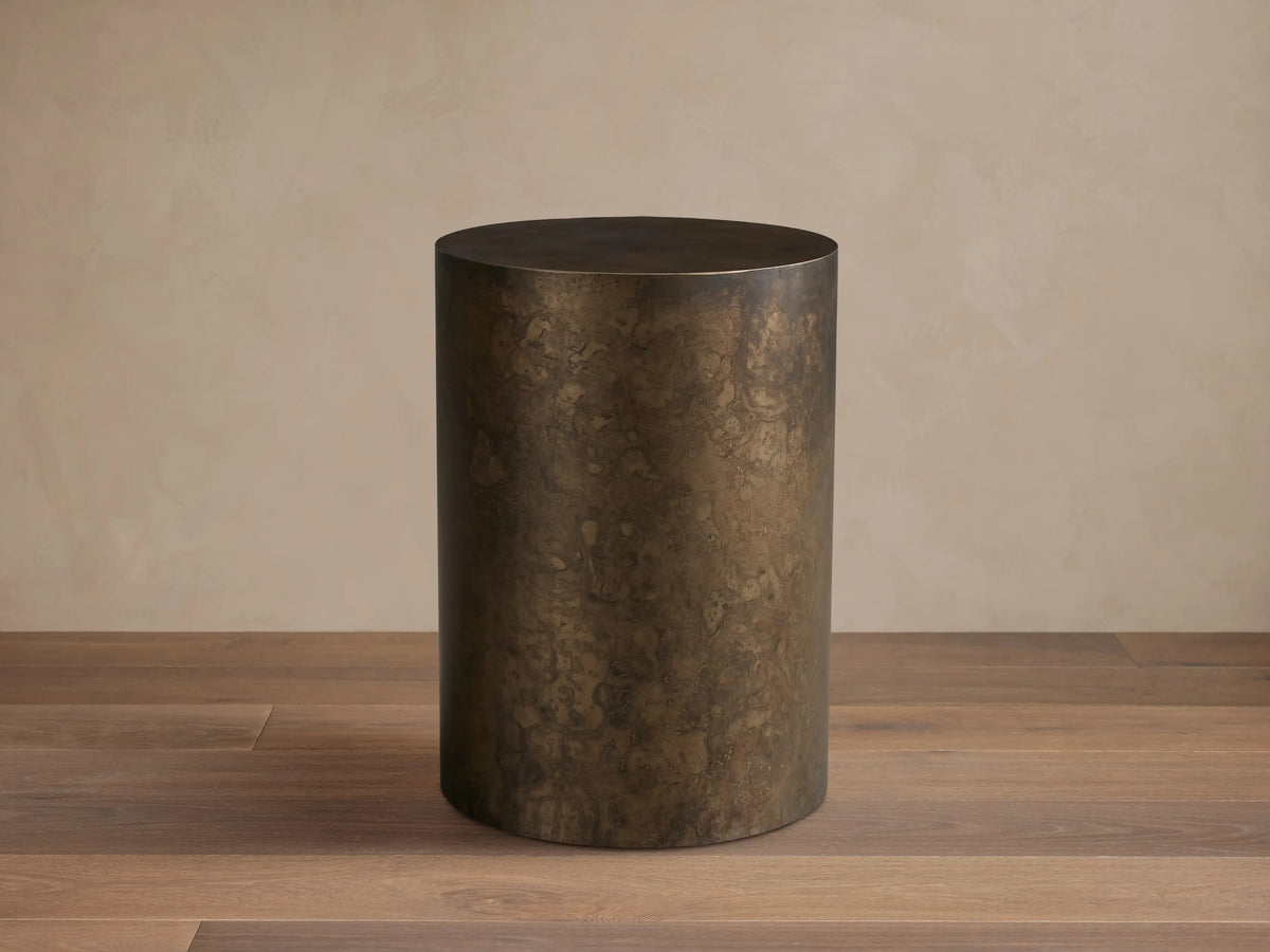 Bruna End Table, cylindrical, dark metal with a warm, weathered patina, on a wooden floor against a neutral wall.