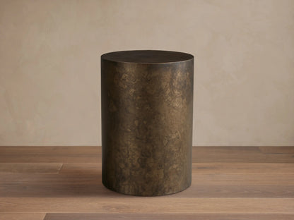 Bruna End Table, cylindrical, dark metal with a warm, weathered patina, on a wooden floor against a neutral wall.