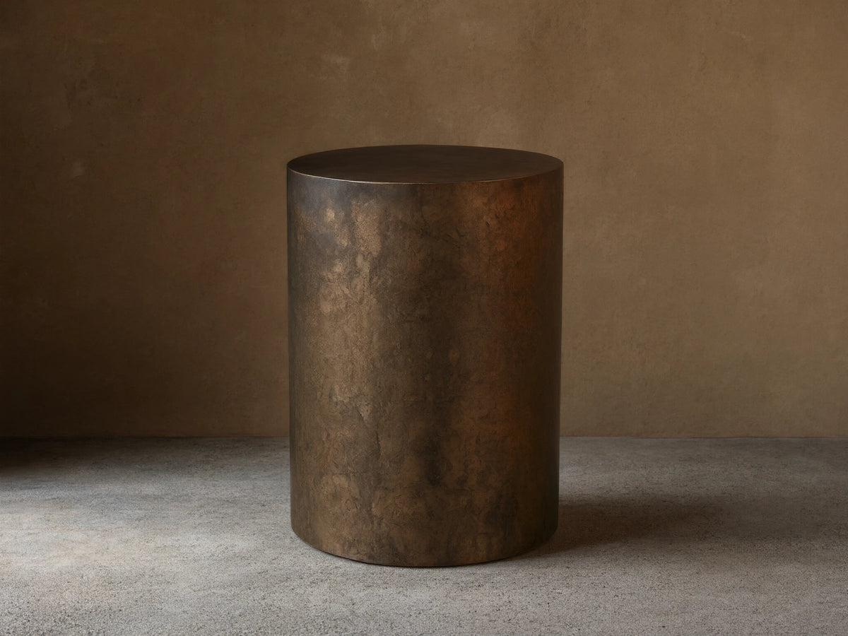 Bruna End Table, cylindrical, dark metal with a warm, weathered patina, on a wooden floor against a neutral wall.