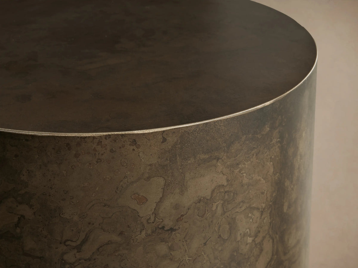 Bruna End Table, cylindrical, dark metal with a warm, weathered patina, on a wooden floor against a neutral wall.