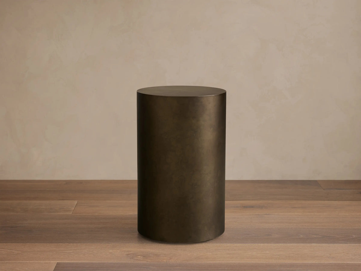 Front view of the dark bronze cylindrical Bruna Pedestal with a weathered metallic patina, on a wooden floor.