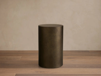 Front view of the dark bronze cylindrical Bruna Pedestal with a weathered metallic patina, on a wooden floor.
