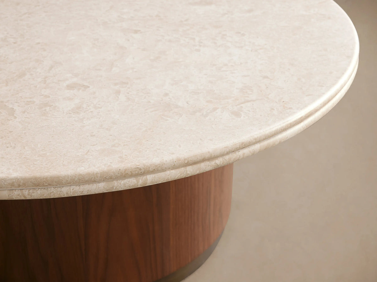 Caden Round Coffee Table with a cream stone top, walnut wood veneer cylindrical base, and antique bronze plinth on wood floor.