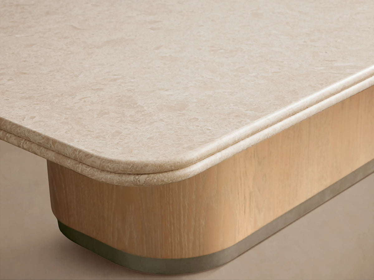 Caden Coffee Table with a cream stone top, sand oak wood veneer base, and brushed nickel plinth on a wooden floor.