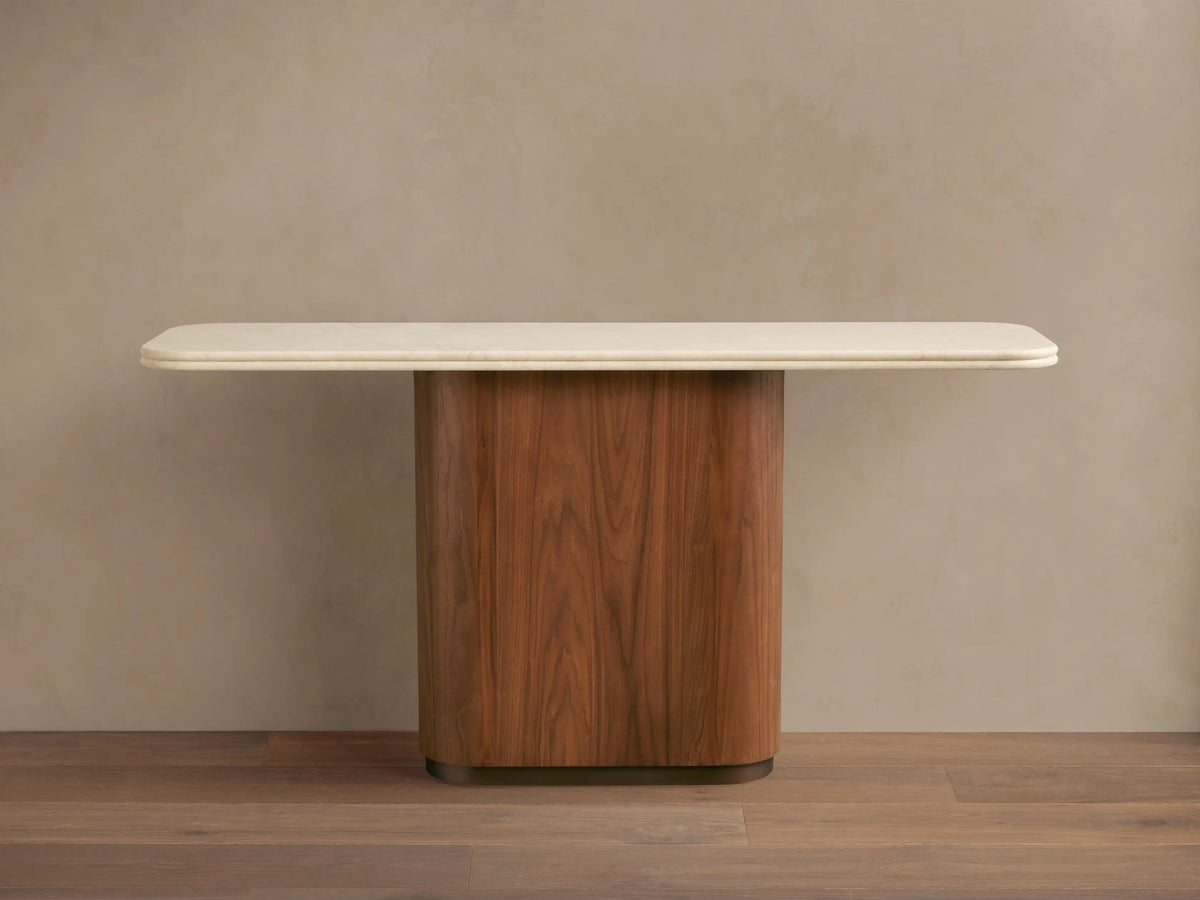 Caden Console Table with cream stone top and rounded walnut wood veneer base. A dark bowl and vase with dried plants sit on top.