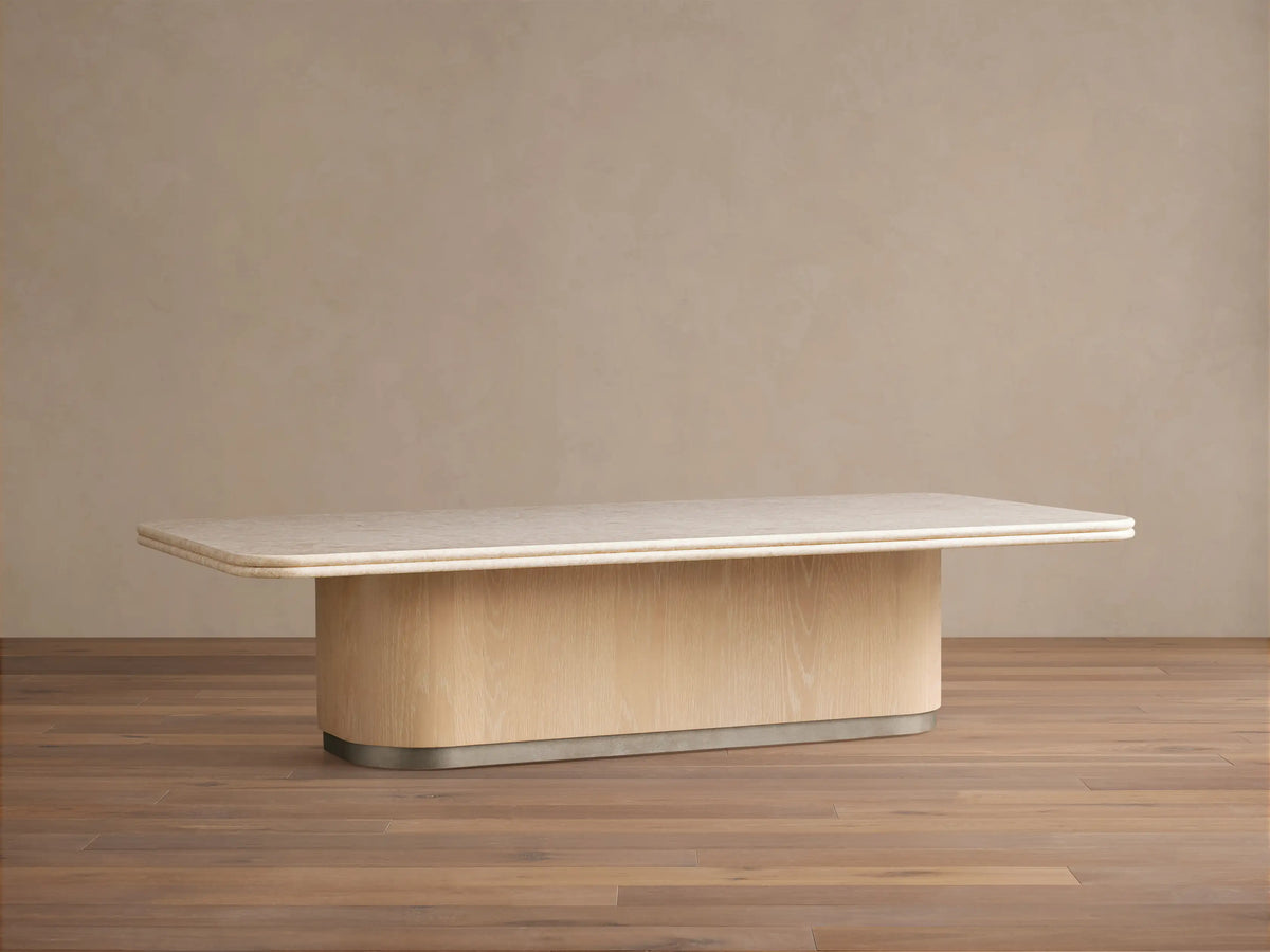 Caden Coffee Table with light cream stone top, sand oak veneer curved base, and brushed nickel plinth on wood floor.
