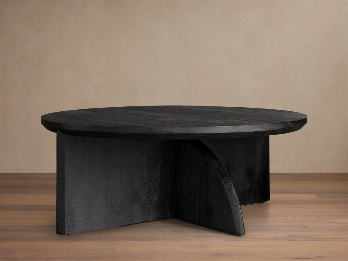 Calderone Coffee Table with a round top in matte black finish, featuring flat and curved intersecting solid wood legs on a wood floor.