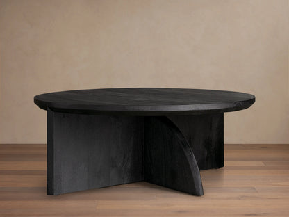 Calderone Coffee Table with a round top in matte black finish, featuring flat and curved intersecting solid wood legs on a wood floor.