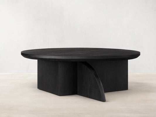 Calderone Coffee Table with a round top in matte black finish, featuring flat and curved intersecting solid wood legs on a wood floor.