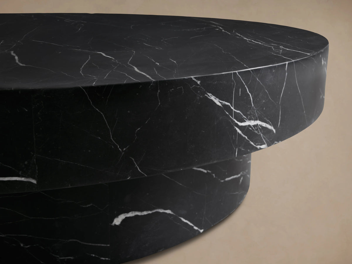 Cinzia Stone Round Coffee Table in black marble with white veining, featuring a low-profile, plinth-style base on a wooden floor.