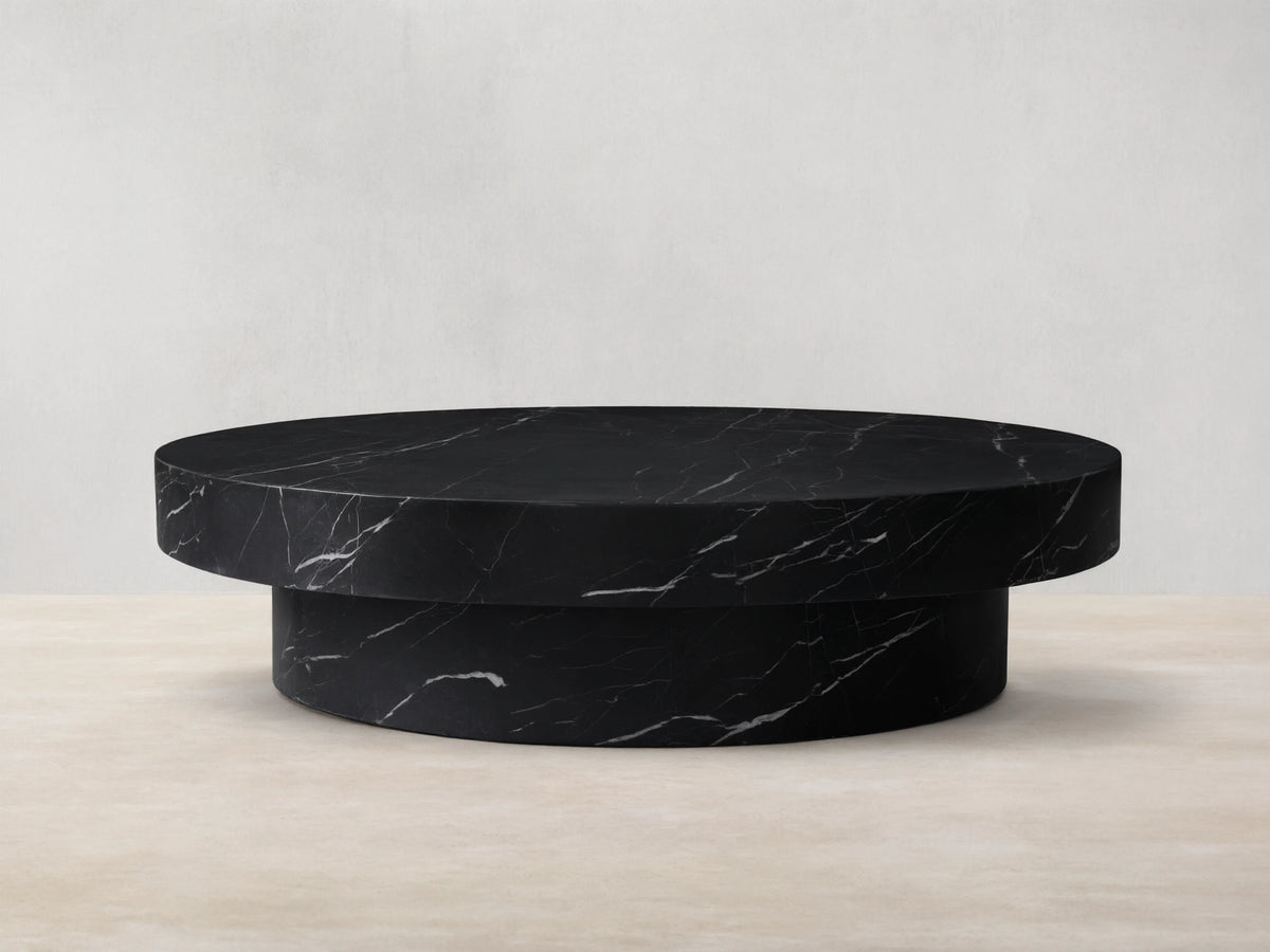 Cinzia black marble round coffee table with prominent white veining, low-profile plinth base, on a wooden floor.