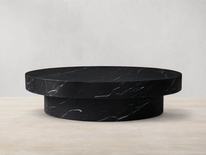 Cinzia black marble round coffee table with prominent white veining, low-profile plinth base, on a wooden floor.