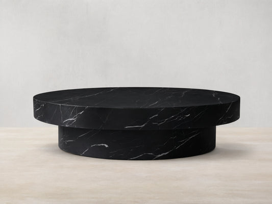 Cinzia black marble round coffee table with prominent white veining, low-profile plinth base, on a wooden floor.