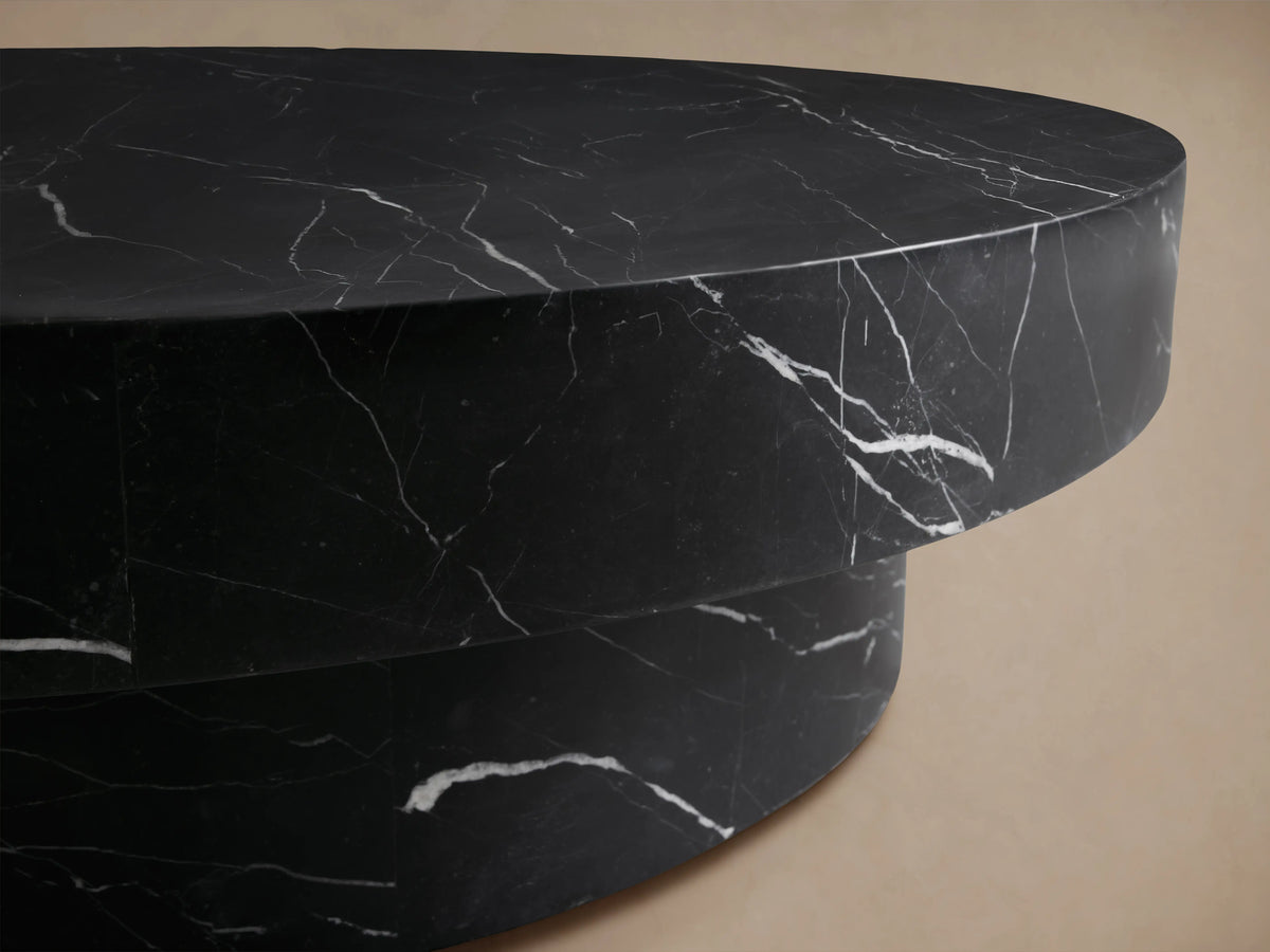 Cinzia Stone Round Coffee Table with black marble finish, low-profile silhouette, plinth-style base on wooden floor.