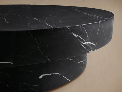 Cinzia Stone Round Coffee Table with black marble finish, low-profile silhouette, plinth-style base on wooden floor.