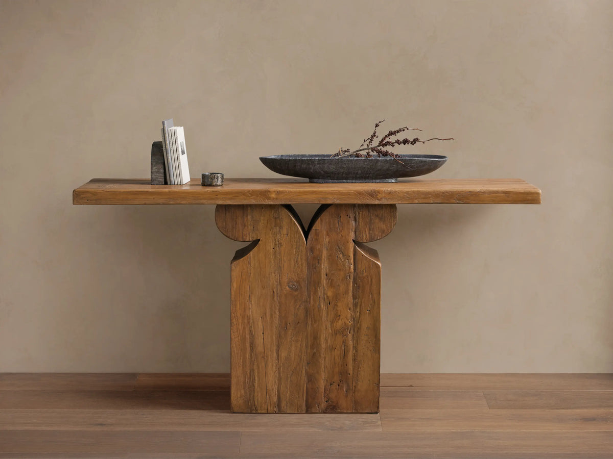 Wooden Dumai Console Table with a slab top and decorative pedestal base. On top are books, a small candle, and a black bowl with dried branches.