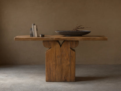 Wooden Dumai Console Table with a slab top and decorative pedestal base. On top are books, a small candle, and a black bowl with dried branches.