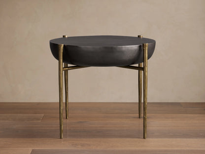Donovan End Table with a round, black nickel textured surface and antique brass legs on a wooden floor background.