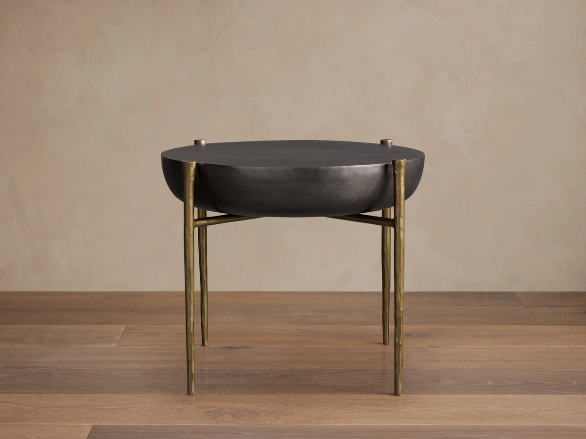 Donovan End Table with a round black nickel surface and slender antique brass legs, set on a wooden floor against a beige wall.