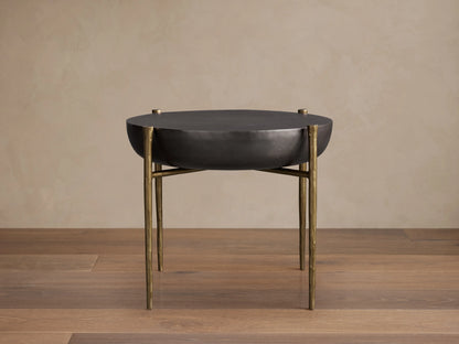 Donovan End Table with a round black nickel surface and slender antique brass legs, set on a wooden floor against a beige wall.