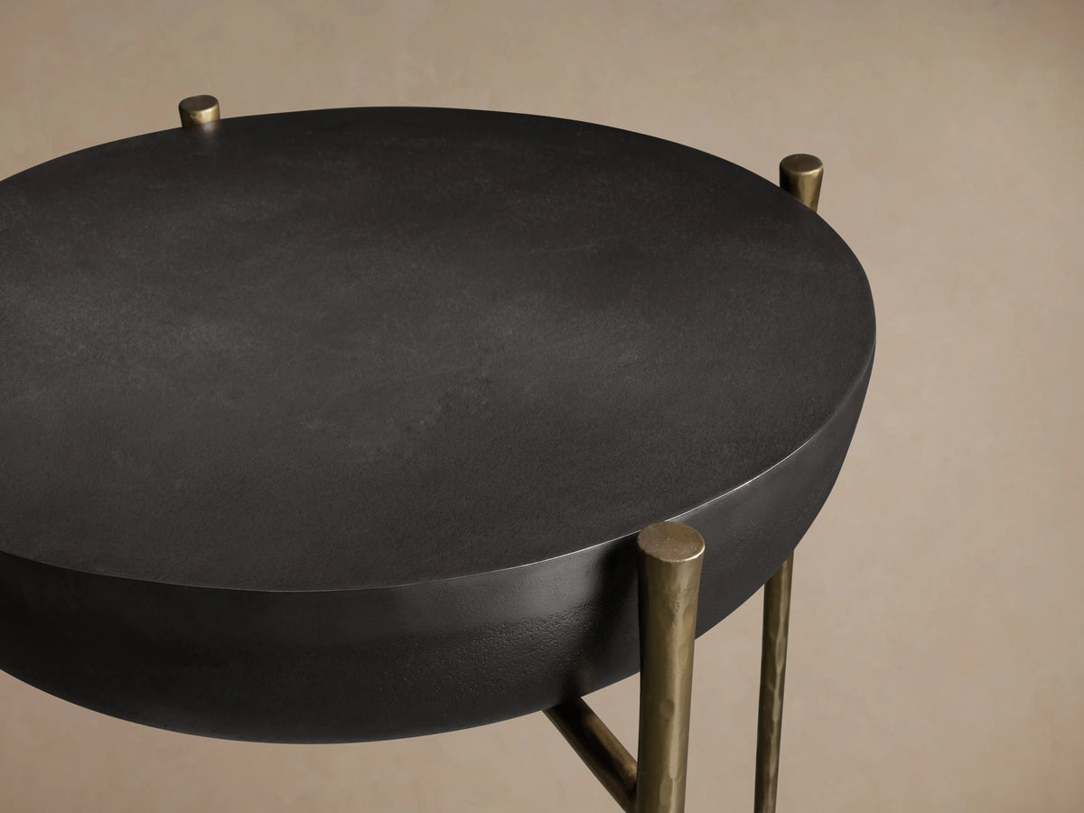 Donovan End Table with a round, black nickel textured surface, supported by slender, antique brass legs on a beige background.