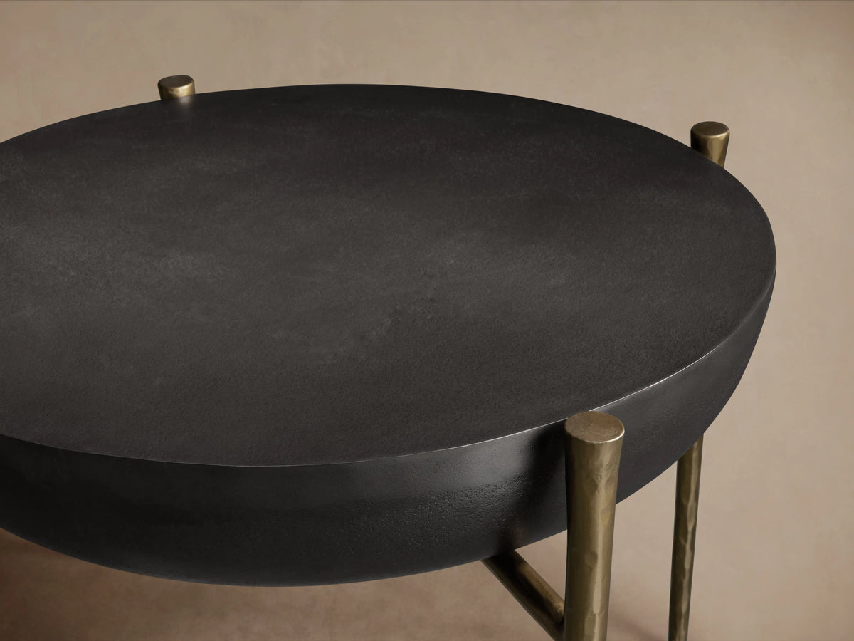 Donovan End Table with a round, black nickel textured surface and antique brass legs on a wooden floor background.