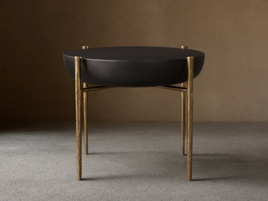 Donovan End Table with a round, black nickel textured surface and antique brass legs on a wooden floor background.