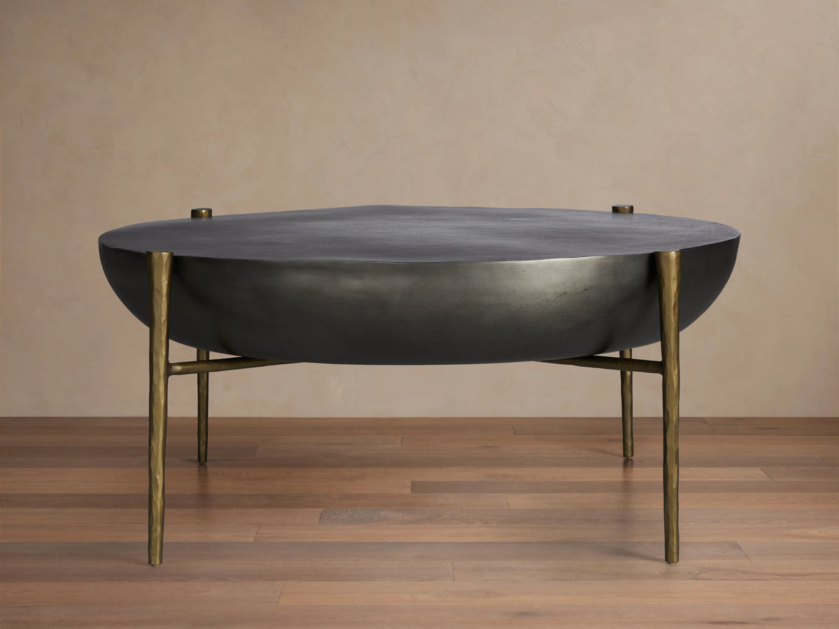 Donovan Coffee Table in black nickel finish with round, textured cast aluminum top, antique brass legs, on wooden floor.