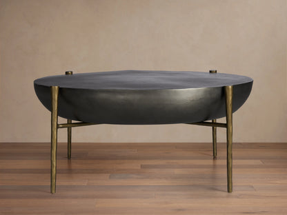 Donovan Coffee Table in black nickel finish with round, textured cast aluminum top, antique brass legs, on wooden floor.