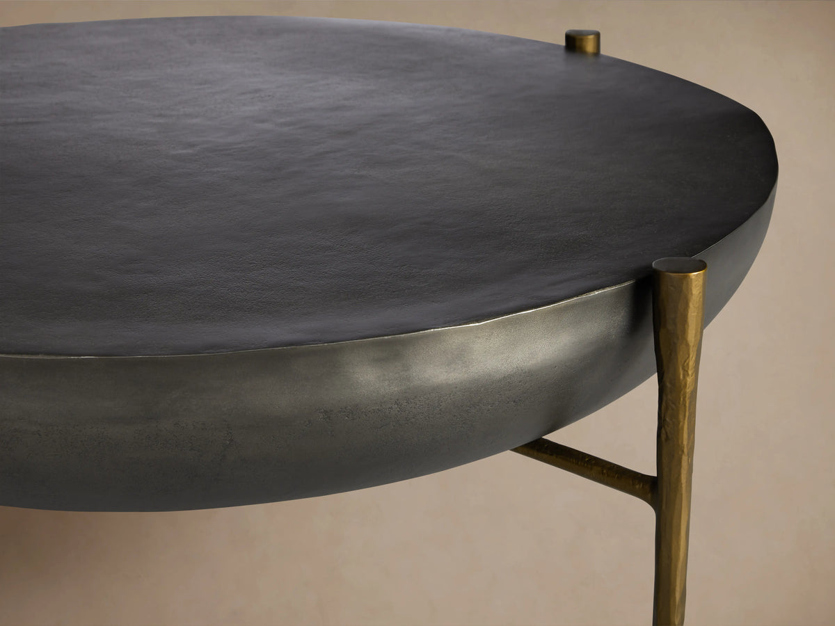 Donovan Coffee Table in black nickel finish with round, textured cast aluminum top, antique brass legs, on wooden floor.