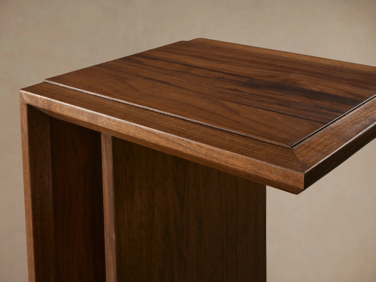 Elien C Table in a warm wood finish, side view showing its C-shaped design on a wooden floor, against a beige wall.