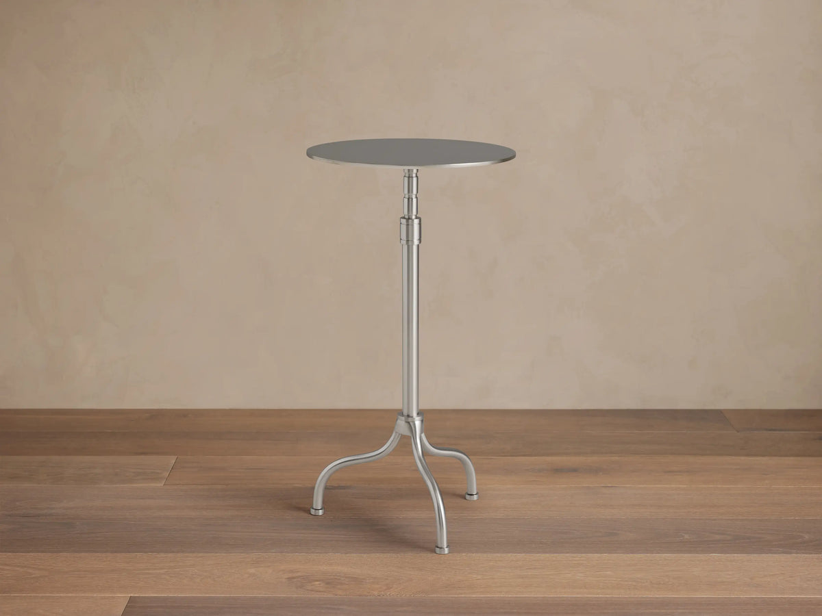 Ferdinand Martini Table with a sleek round metal top and a slim central stem on three curved legs, set against a wooden floor.