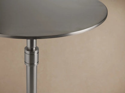 Ferdinand Martini Table with a sleek round metal top and a slim central stem on three curved legs, set against a wooden floor.